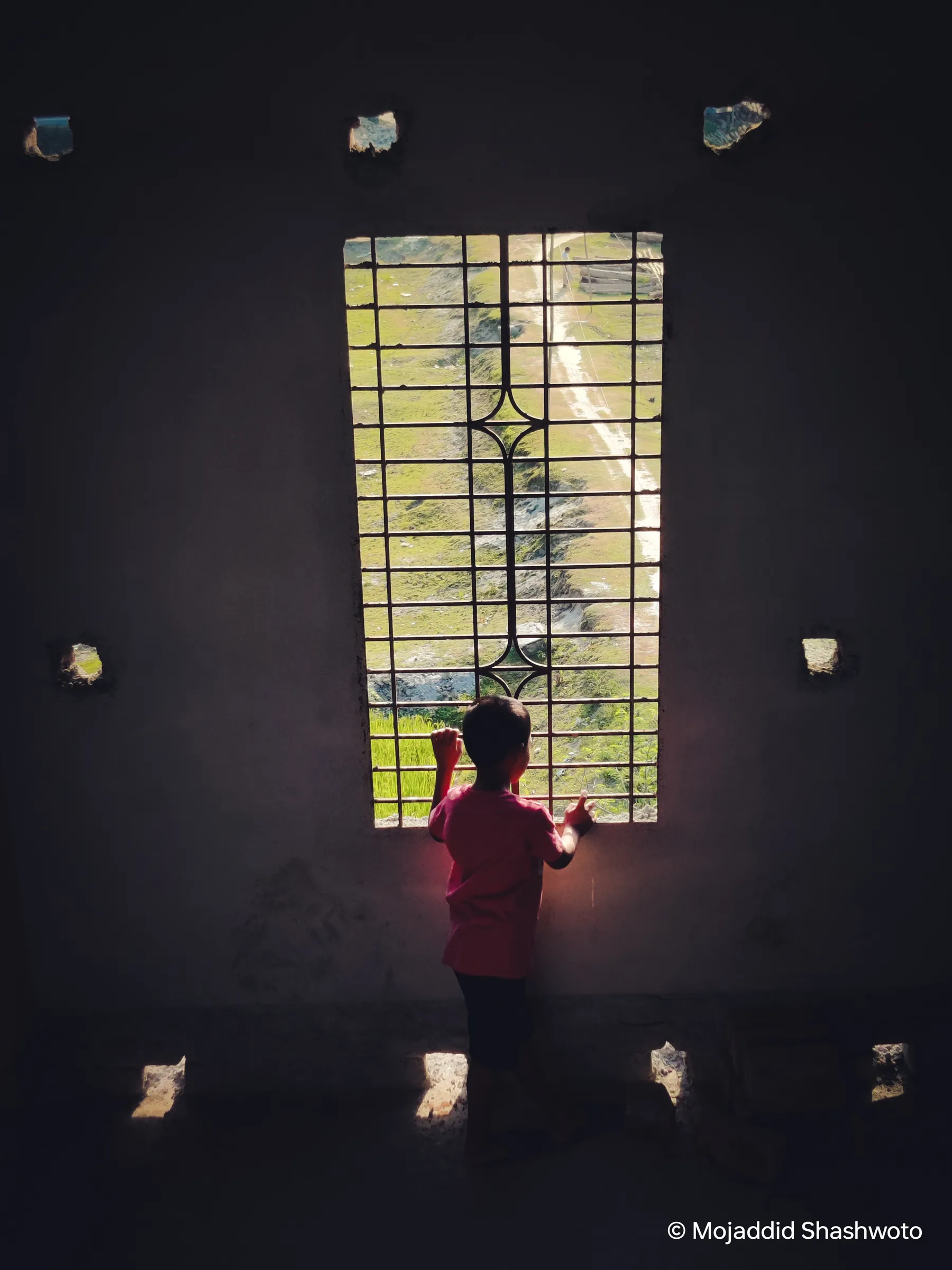 Child silhouetted at a barred window overlooking a sunlit green hillside