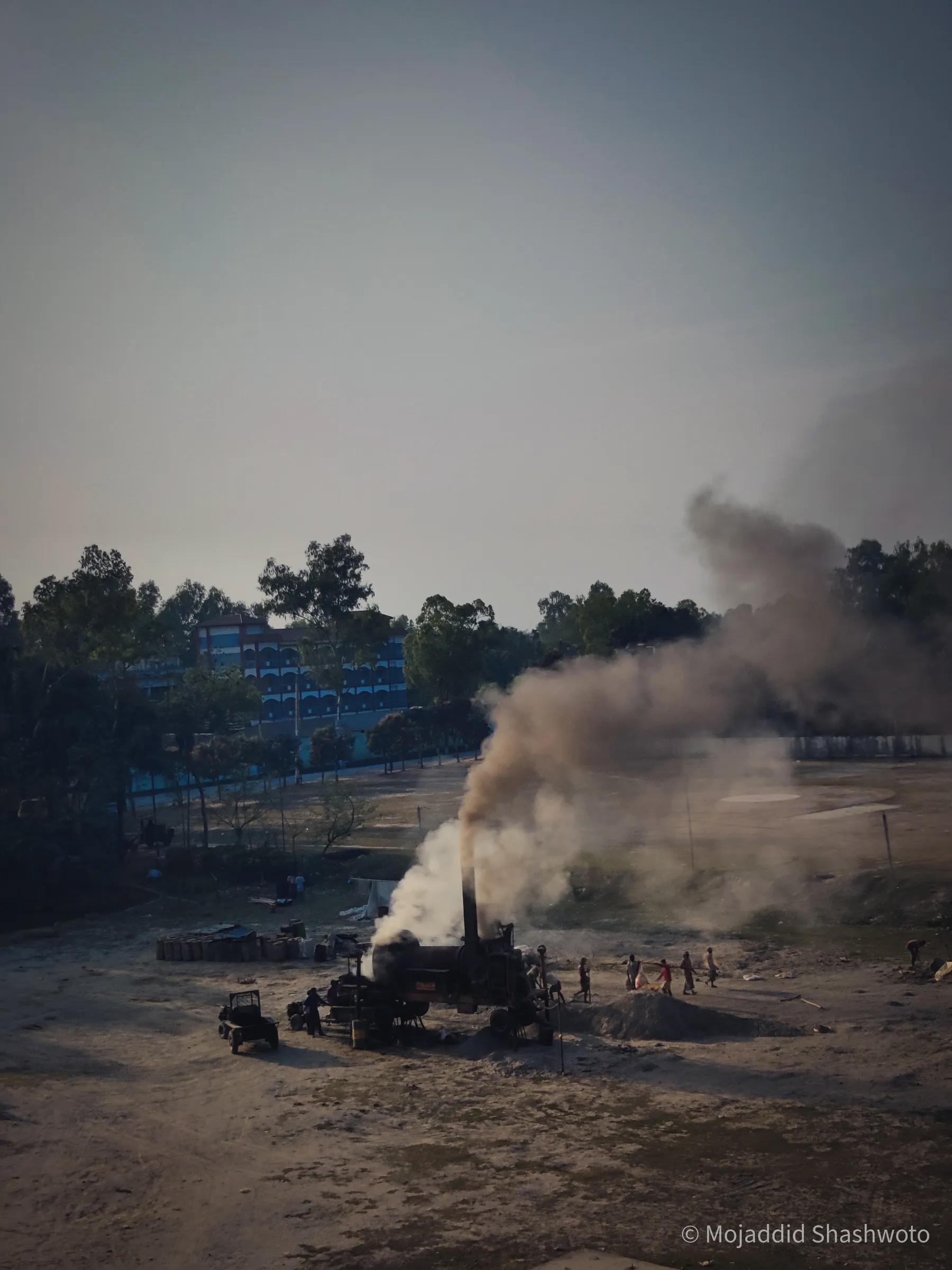 Roadwork asphalt mixer billowing dark smoke across an open clearing with workers