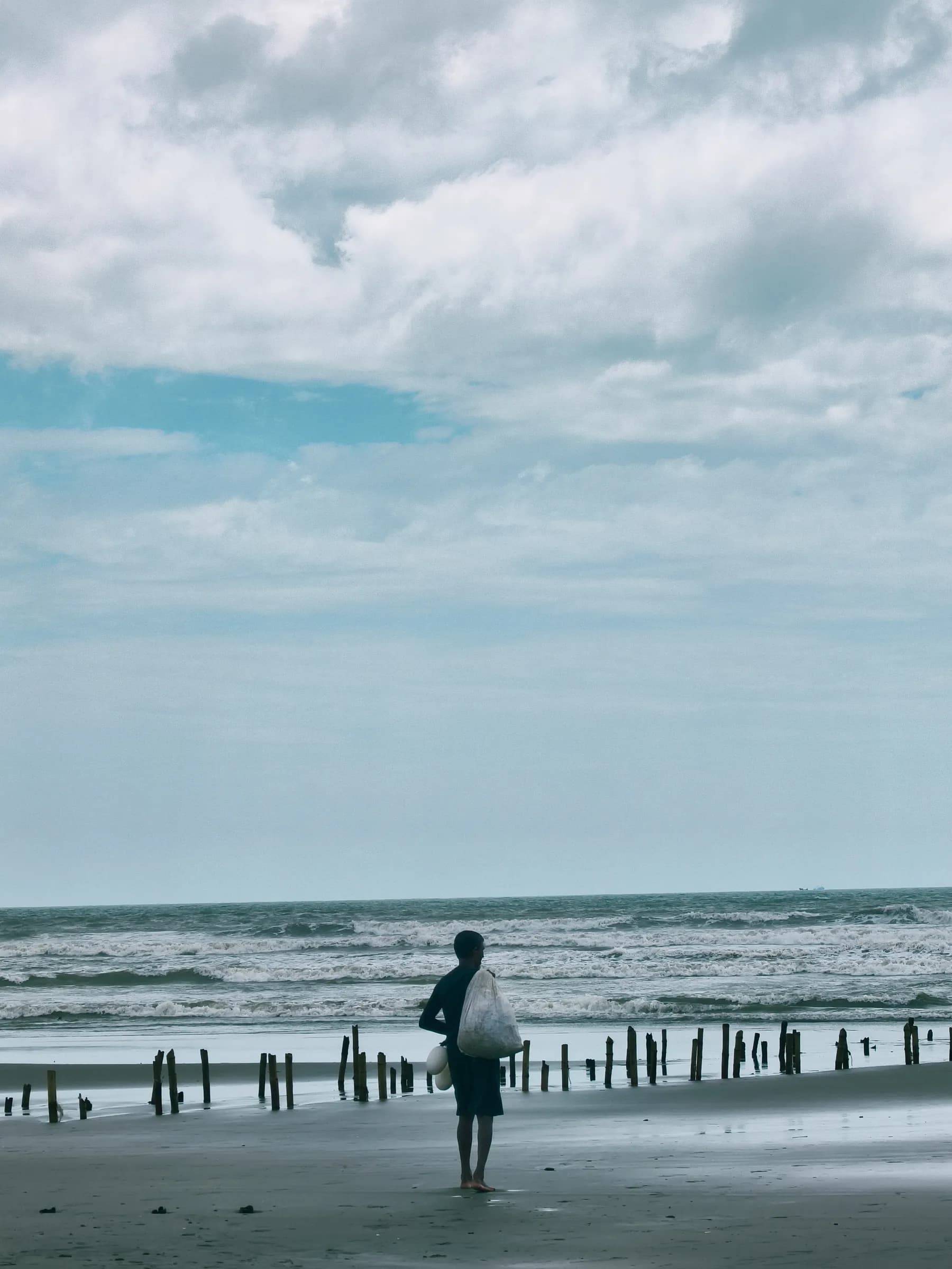Solitary figure walking across sandy beach towards wooden poles in breaking waves